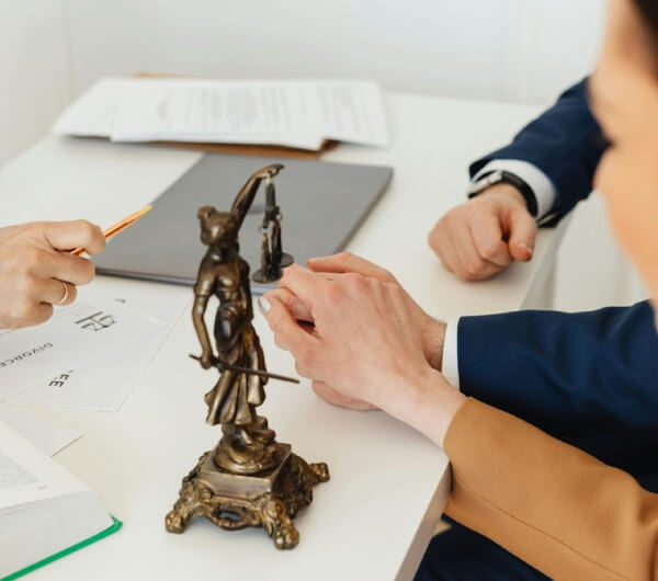 A man and woman holding hand while talking to a lawyer who has a statue holding scales on his desk.