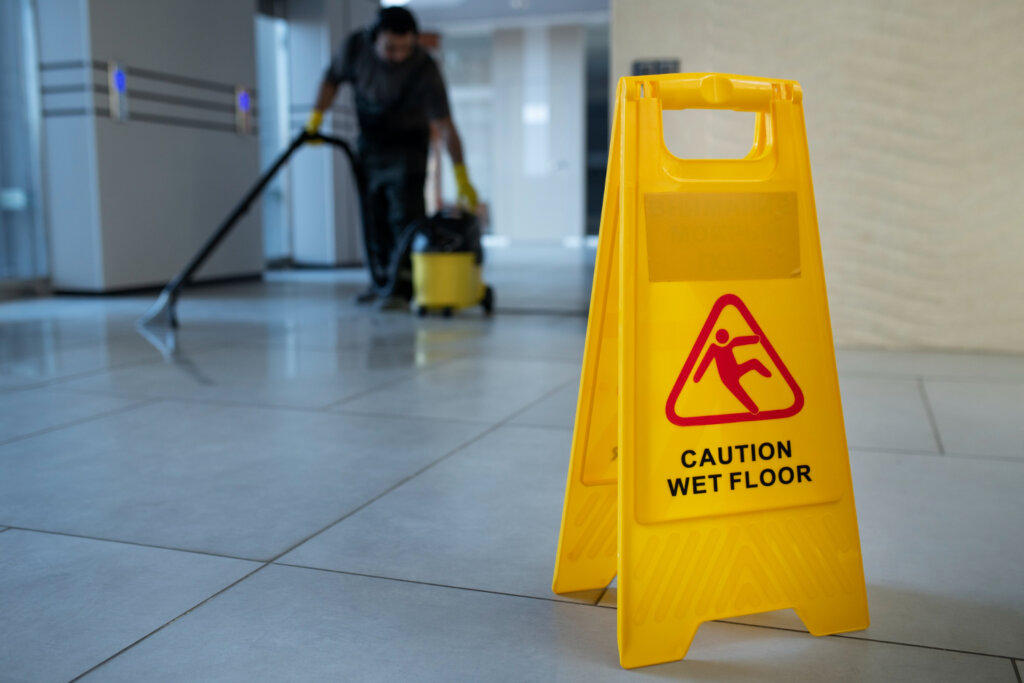a yellow wet floor sign with a janitor cleaning a hallway in the background